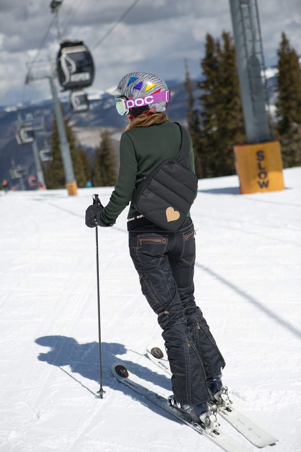 Skier wearing the black puffer sling backpack with gold heart on a snowy slope beside a gondola lift