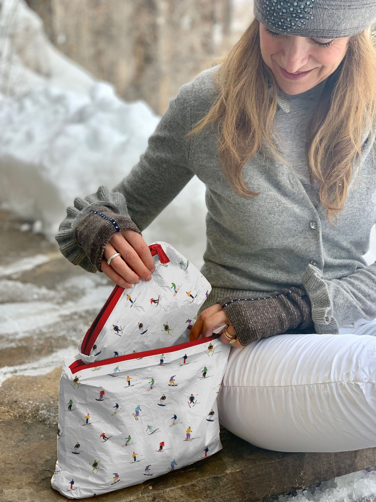 Woman sitting outside in snowy environment with two coordinating pouches, one mini and one medium with dancing skiers print on each. 