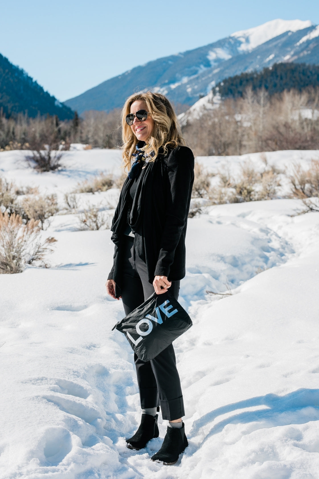 Woman in a black outfit and boots walking through snow, carrying the Medium Pouch in Black with Silver LOVE