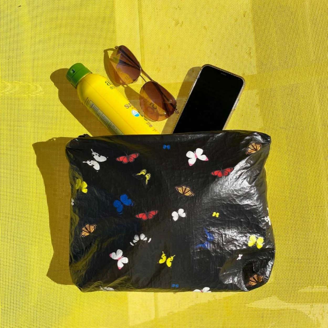 Black pouch with colorful butterflies holding sunblock, sunglasses, and cell phone on yellow background