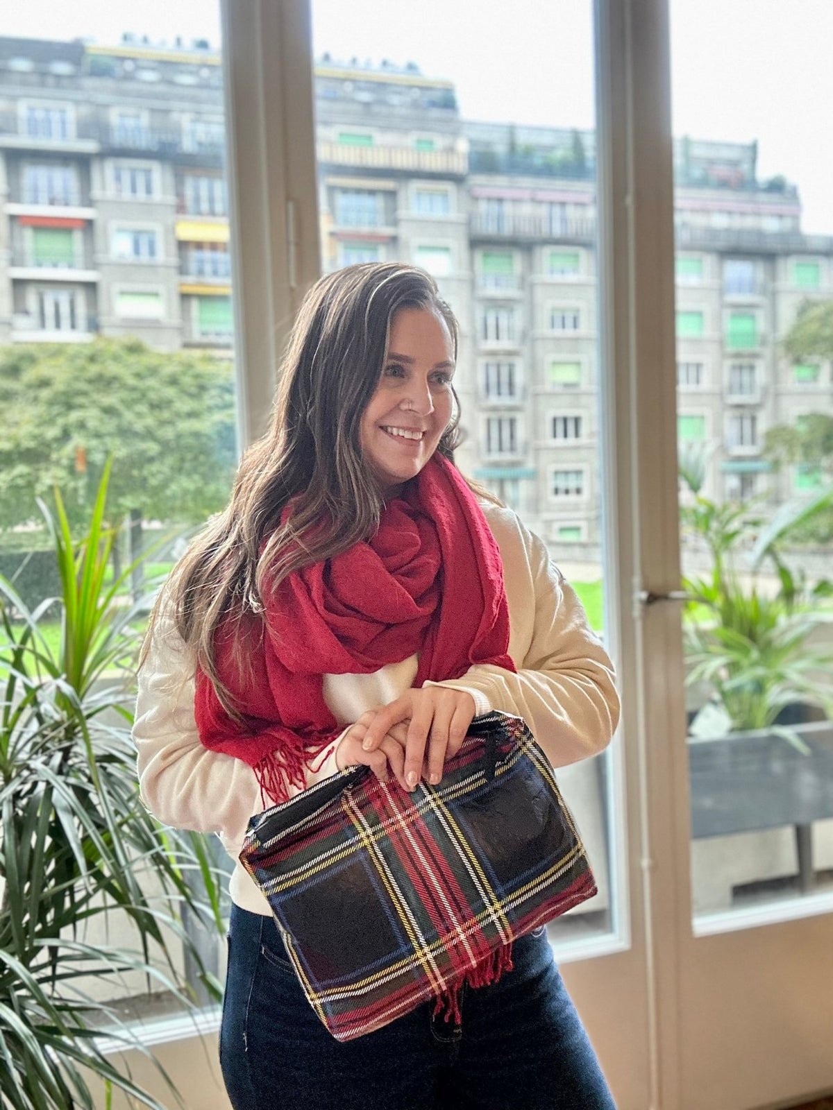 Woman in red scarf smiling holding tartan plaid medium pouch with zipper against window background.