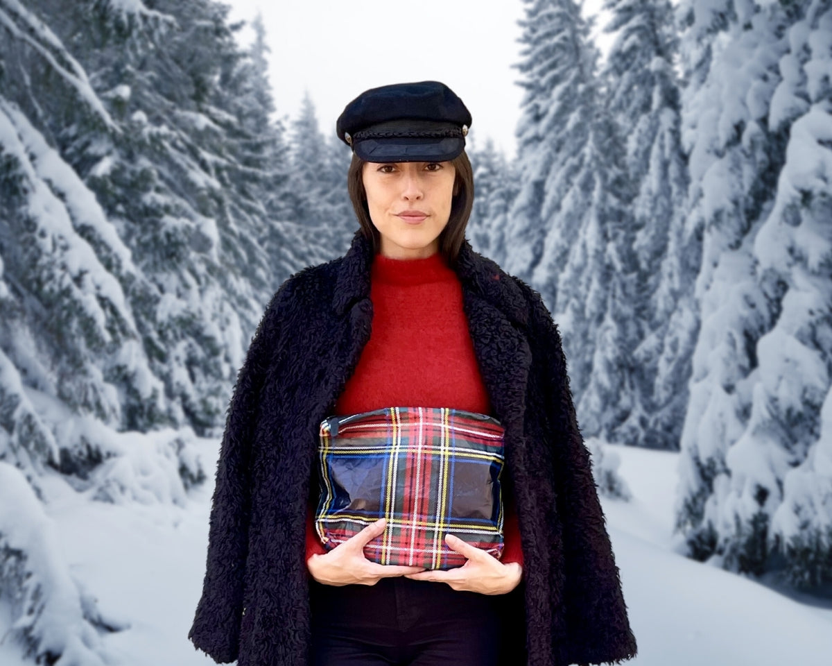 Woman in red top and fur coat holding Medium zipper pouch in tartan plaid pattern against snowy tree background. 