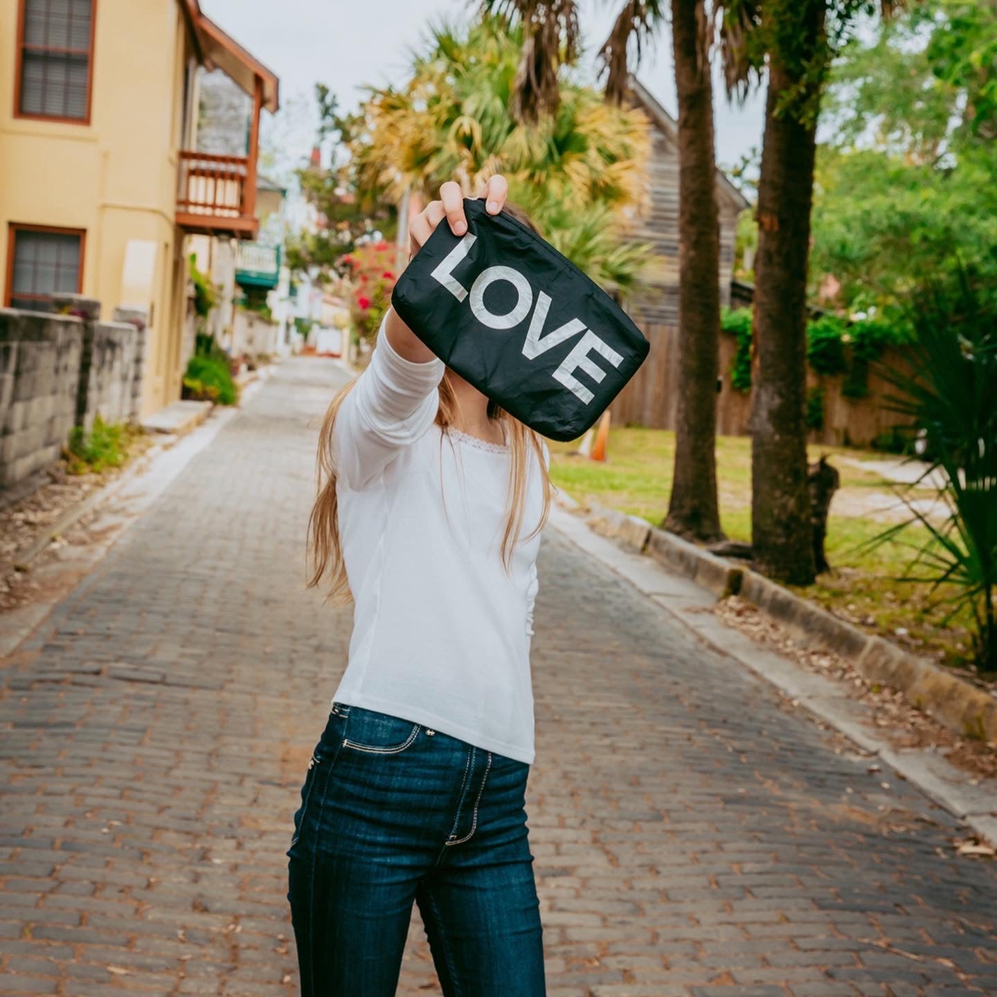 Woman on a cobblestone street holding up a black "LOVE" pouch on a tree-lined lane.