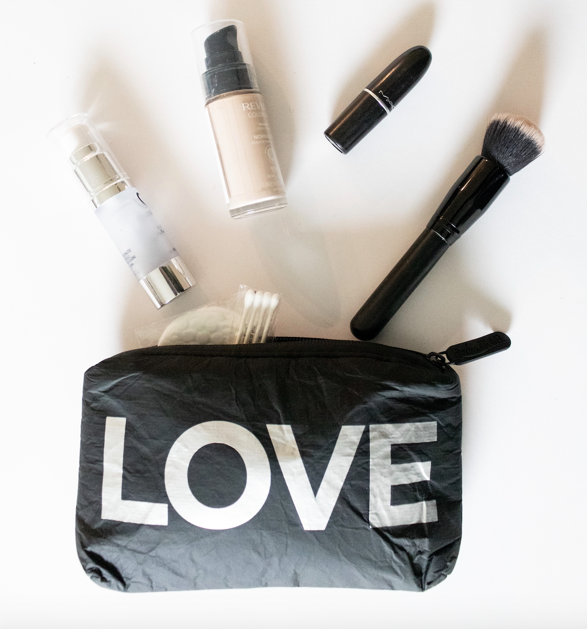 Black "LOVE" pouch flat lay spilling out makeup essentials — foundation, lipstick, brush, and skincare products.