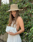 Woman standing near greenery wearing white dress and sunhat with bee pouch