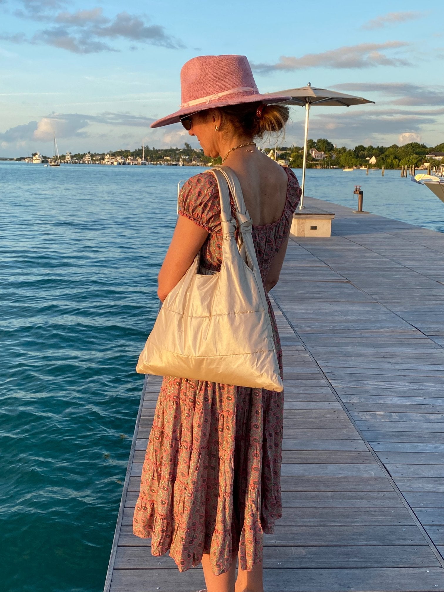 Woman in pink hat and dress standing on ocean dock with shimmer beige shoulder tote