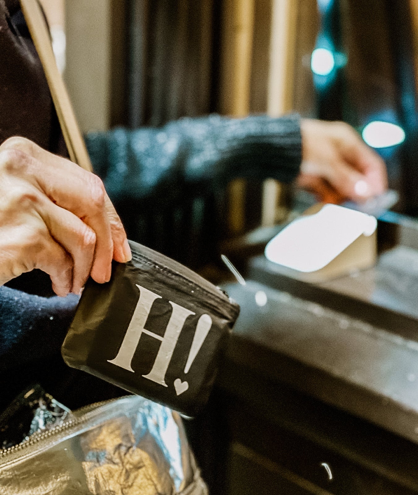 Hands opening the black H! Card Pouch on a wooden counter with a mirror and makeup sponge nearby