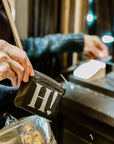 Hands opening the black H! Card Pouch on a wooden counter with a mirror and makeup sponge nearby