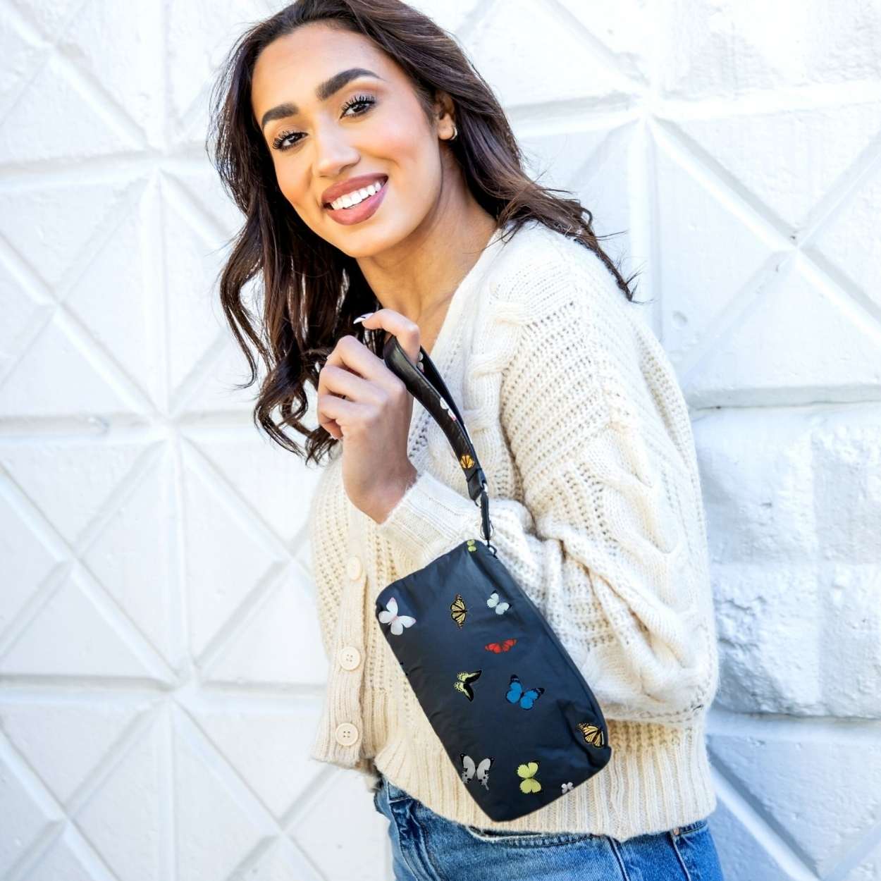 Woman in cream sweater and jeans holding black wristlet with butterflies against a white wall