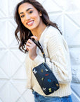 Woman in cream sweater and jeans holding black wristlet with butterflies against a white wall